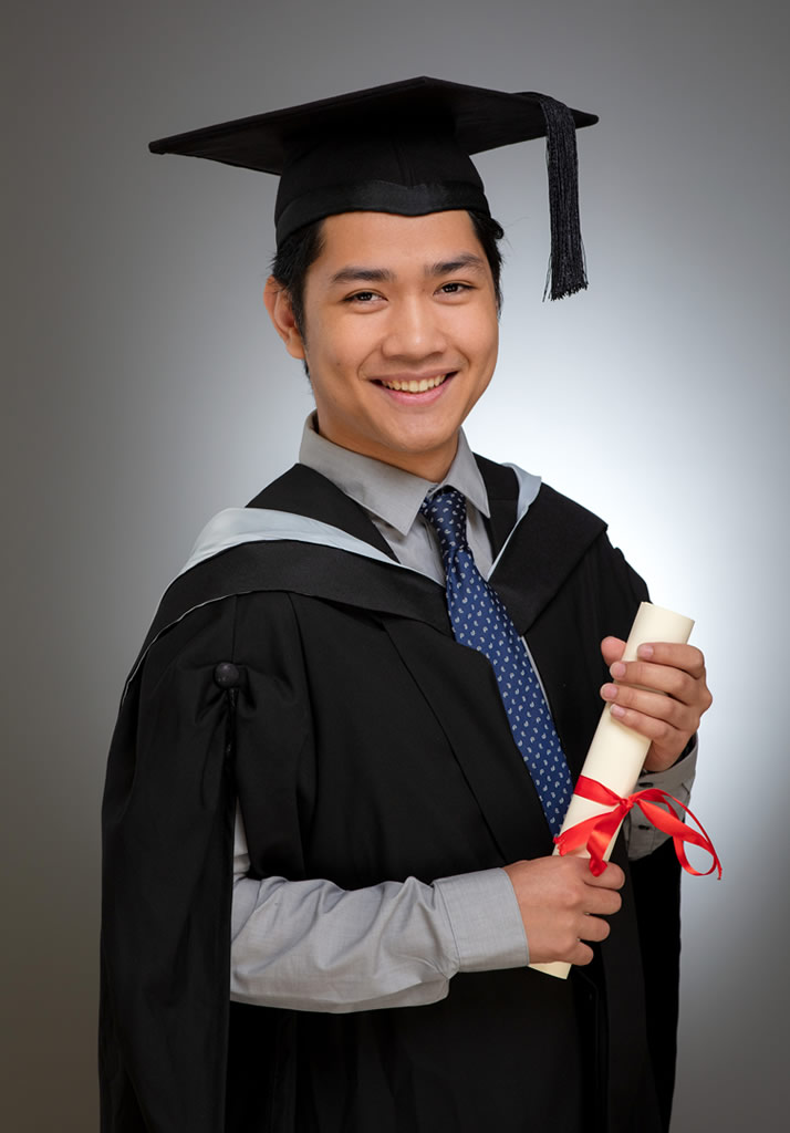 Studio graduation portrait by Udayaa Studio — graduate in cap and gown holding diploma scroll, clean grey backdrop