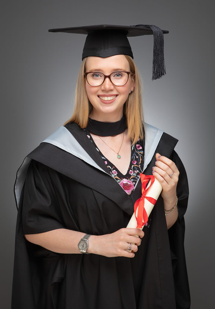 Studio graduation portrait by Udayaa Studio — graduate in academic gown with diploma, clean studio grey backdrop