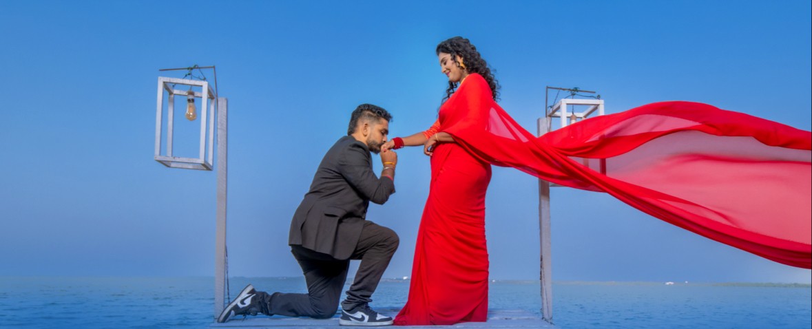 Pre-wedding outdoor shoot by Udayaa Studio — couple on a coastal jetty, scarlet saree against open blue sky